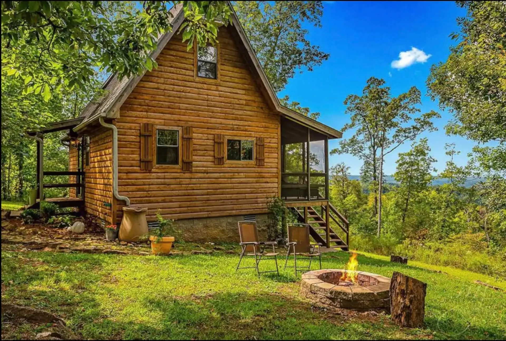 A Still Point Cabin, a two-storey Ozark log cabin, with a stone fire pit and chairs on the lawn and the valley falling away beyond