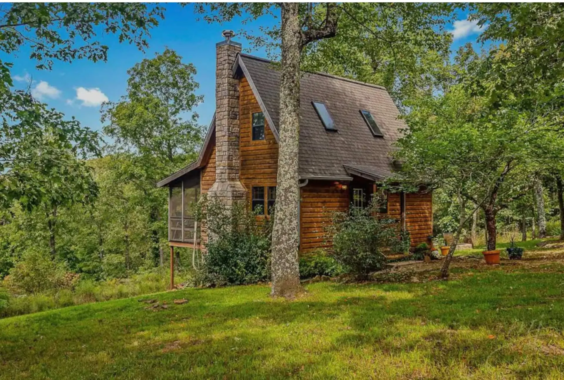 The log cabin seen from the meadow, with its tall stone chimney, dormer windows and screened porch surrounded by Ozark hardwoods