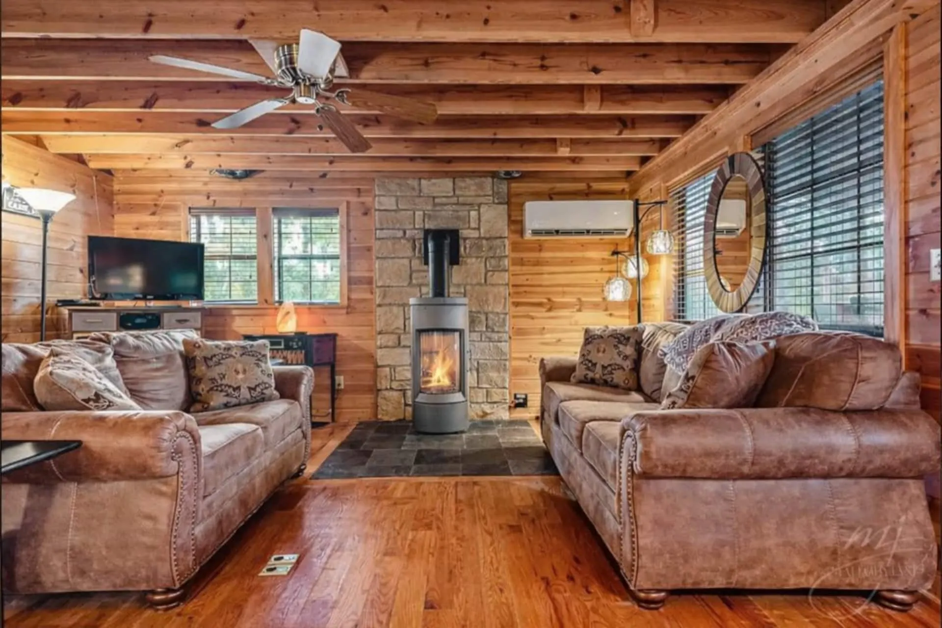 Living room with a gas fireplace set into a stone surround, two leather sofas, a wall-mounted TV and a vaulted pine ceiling